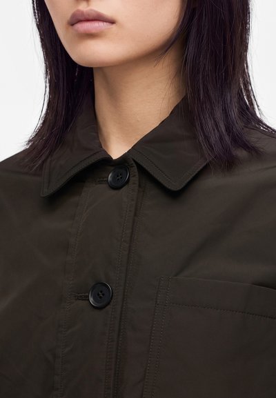 Femme aux cheveux raides et foncés portant une veste marron foncé boutonnée avec un col et une poche poitrine, regardant vers l'avant avec une expression neutre.