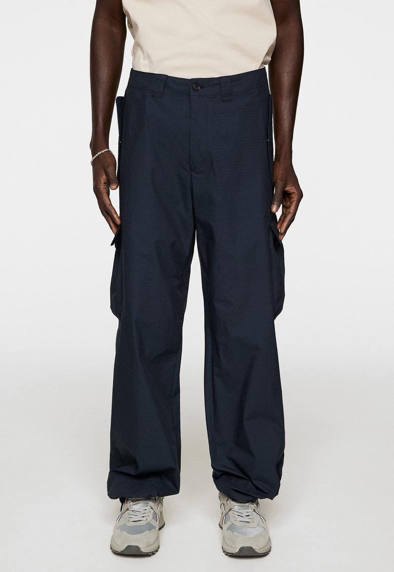 Marinefarbene Cargo-Hosen mit lockerer Passform, strukturiertem Stoff, Seitentaschen und Knopfverschluss, kombiniert mit hellgrauen Sneakers.