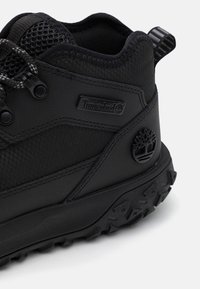 Black hiking boot featuring mesh panels, textured fabric, round laces, and a rubber sole with patterned tread. Timberland logo on the side.