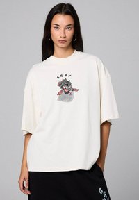 Camiseta de gran tamaño en color crema de algodón con un gráfico colorido de un rostro y el texto "GRMY" en el pecho. Manga corta y corte relajado.