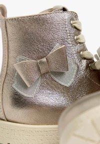 Botte métallisée rose gold avec une finition texturée, ornée d'un grand nœud en tissu et de lacets beige sur une semelle claire.