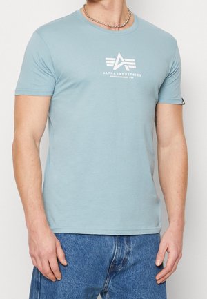 Mężczyzna w jasnoniebieskiej koszulce Alpha Industries i niebieskich dżinsach, pokazujący przednią górną część ciała i ramiona.