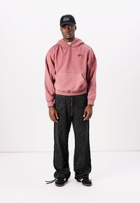 Haut à capuche rose surdimensionné avec logo, pantalon noir texturé avec motif blanc, et casquette noire avec texte blanc, porté avec des baskets noires.