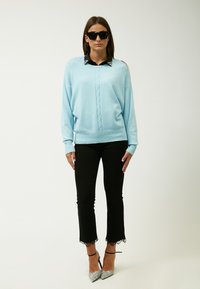 Hellblauer Pullover mit einem Kabelstrickdesign, kombiniert mit schwarzen Hosen mit Spitze. Das Modell trägt schwarze Sonnenbrillen und silberne High Heels.