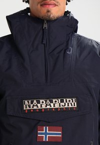 Die marineblaue Pulloverjacke verfügt über einen hohen Kragen, einen Reißverschluss, eine große frontale Tasche und markierte Patches, darunter ein Detail der norwegischen Flagge.