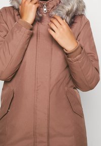 Brązowa ocieplana kurtka z kapturem z sztucznego futra, z zasuwakiem z przodu, kieszeniami bocznymi i ściągaczami na mankietach, o gładkiej teksturze i lekko błyszczącym wykończeniu.