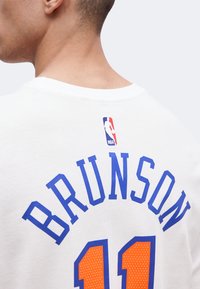 Πλάτη ενός ατόμου που φορά λευκή φανέλα NBA με το όνομα "BRUNSON" και τον αριθμό "11" σε μπλε και πορτοκαλί χρώματα.