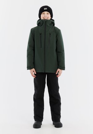 Groene waterdichte jas met een hoge kraag, twee ritszakken op de borst en zwarte broek. Het model draagt een zwarte beanie en instap schoenen.
