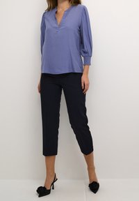 Chemisier léger bleu à manches longues avec un col en V et des poignets élastiqués, associé à un pantalon noir coupé et des chaussures à talons noires.