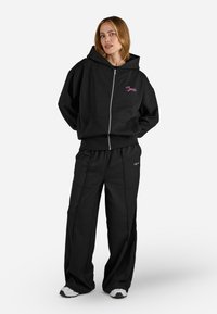 Felpa nera con zip, cappuccio e tasche anteriori, abbinata a pantaloni neri larghi. Entrambi presentano delicati dettagli ricamati in rosa.