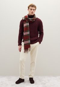 Bordeauxroter Strickpullover mit Zopfmuster, kariertes Halstuch in Rot, Grün und Weiß, cremefarbene Hose und dunkelbraune Wildlederschuhe.