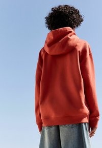 Burnt orange oversized hoodie with a drawstring hood, ribbed cuffs, and hem, paired with light denim shorts against a clear sky.
