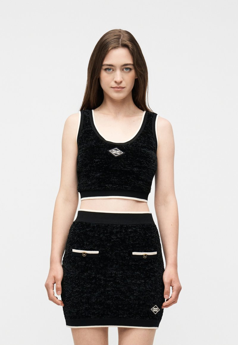 Νεαρή γυναίκα που φοράει μαύρο υφασμάτινο αμάνικο crop top και το αντίστοιχο φούστα με λευκή διακόσμηση και μικρές τσέπες, στέκεται μπροστά σε απλό φόντο.