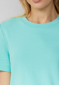 Un t-shirt en coton turquoise avec un col rond, des manches courtes et une texture lisse, présentant des détails de couture le long des coutures.