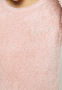 Ljusrosa, fluffig tröja med rund halsringning och en mjuk textur. Har ordet "GUESS" i glittrande bokstäver på framsidan.