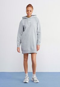 Grijze fleece sweatshirt jurk met een capuchon, voorzien van een voorzak en ribgebreide manchetten. Model draagt lichte sneakers met witte sokken op een blauwe vloer.