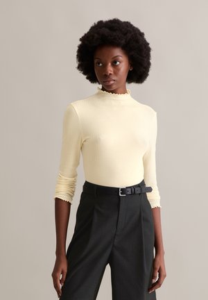 Gelbes, geripptes Langarmoberteil mit einem hohen, gerafften Halsausschnitt, kombiniert mit schwarzen, maßgeschneiderten Hosen, die über einen Gürtel mit silberner Schnalle verfügen.