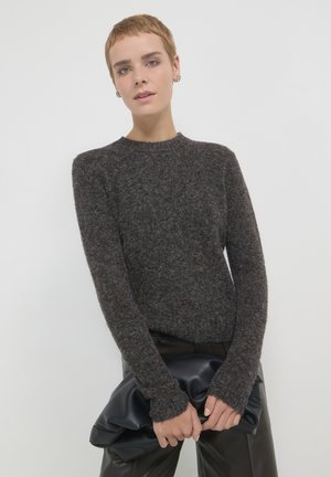 Dunkelgrauer gestrickter Pullover mit rundem Halsausschnitt und langen Ärmeln, mit gerippten Bündchen. Das Model hält eine schwarze Leder-Clutch.