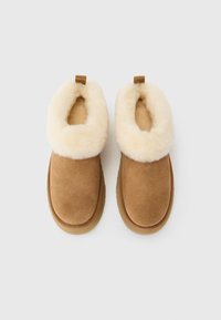 Un paio di pantofole in suede marrone senza lacci con morbido rivestimento color crema e linguette di estrazione su sfondo bianco.