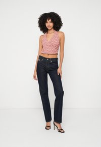 Kvinna med lockigt hår som bär en rosa halter-top, mörkblå jeans och svarta högklackade sandaler, stående mot en vit bakgrund.