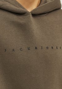 Sudadera marrón hecha de tela suave con un bolsillo canguro. Presenta un logotipo negro elevado de "JACK & JONES" en el pecho.