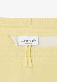 Pantaloni della tuta gialli con vita elasticizzata, dotati di cordoncino e un'etichetta con la scritta "LACOSTE REGULAR FIT." Tessuto di superficie liscia.