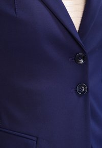 Blazer blu navy realizzato in tessuto liscio, con rever a lancia, due bottoni neri e una tasca singola sul lato sinistro.