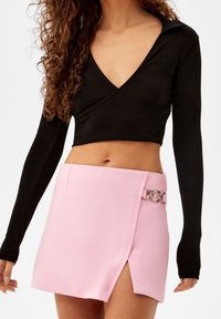 Black long-sleeve, cropped top with a V-neck and collar paired with a pink wrap skirt featuring a metallic accent and side slit.