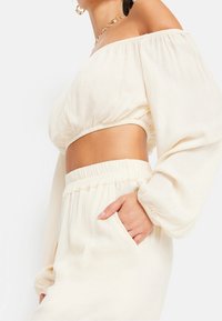 Woman wearing off-shoulder cream crop top with puffed long sleeves and matching high-waisted skirt with hand in pocket.