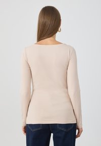 Long-sleeve top in light beige fabric, featuring a square back neckline and a fitted silhouette, paired with dark blue jeans.