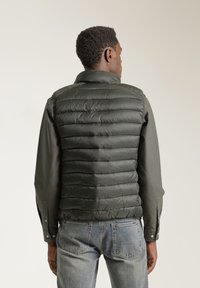 Chaleco acolchado verde oscuro con costuras horizontales, cuello alto y una textura suave, llevado sobre una camisa de manga larga y unos vaqueros.