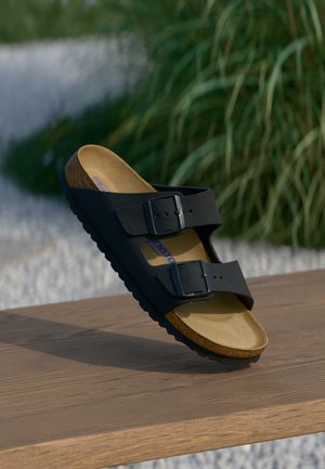 Černé sandály na dvou popruzích s kovovými sponami, korkovou stélkou a texturovanou gumovou podrážkou. Mají hladký svršek a hnědou vnitřní podšívku.