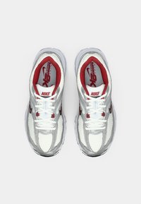 Zapatillas blancas Nike con acentos rojos. Parte superior de malla con patrón texturizado, cuello acolchado y suela redondeada. Cordones blancos, logo visible.