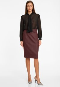 Femme debout portant un chemisier noir transparent à manches longues avec un nœud au cou et une jupe crayon bordeaux taille haute avec de fines rayures.