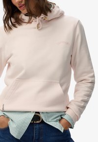 Sudadera con capucha de color rosa claro con herrajes dorados en los cordones, bolsillo frontal y logotipo bordado. Lleva puesta sobre una camisa a rayas en azul verdoso y blanco.
