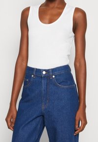 Woman wearing sleeveless white tank top tucked into high-waisted dark blue jeans with front button and pockets visible.