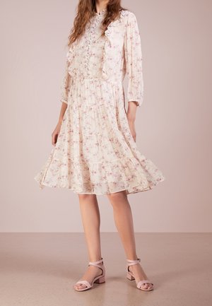 Femme portant une robe fleurie crème mi-longue à manches longues avec des volants, associée à des sandales roses à bride cheville.