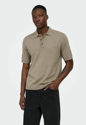 Uomo che indossa una polo beige a maniche corte e pantaloni neri, in piedi con una mano in tasca davanti a uno sfondo neutro.
