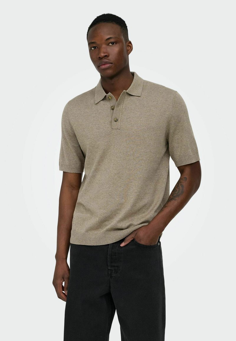Homme portant un polo beige à manches courtes et un pantalon noir, debout avec une main dans la poche sur un fond uni.
