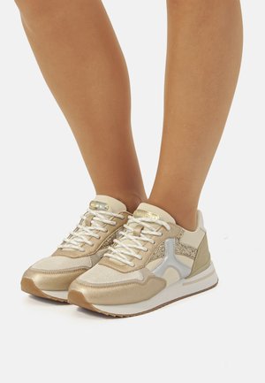 Baskets beiges avec mailles texturées et accents métalliques, dotées de lacets blancs et d'une semelle épaisse et rembourrée. "Conçues avec sens pour le confort."