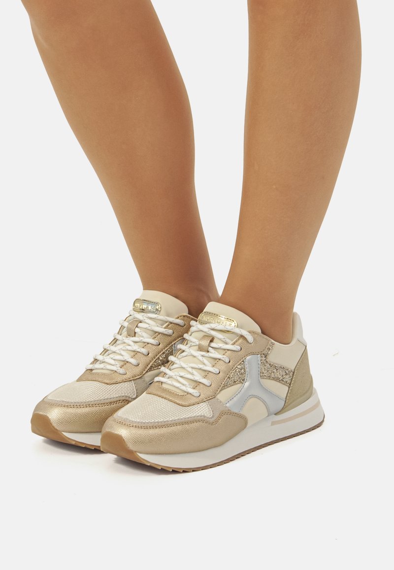 Sneaker beige con mesh testurizzato e dettagli metallici, dotate di lacci bianchi e una suola spessa e imbottita. "Progettate con senso per il comfort."