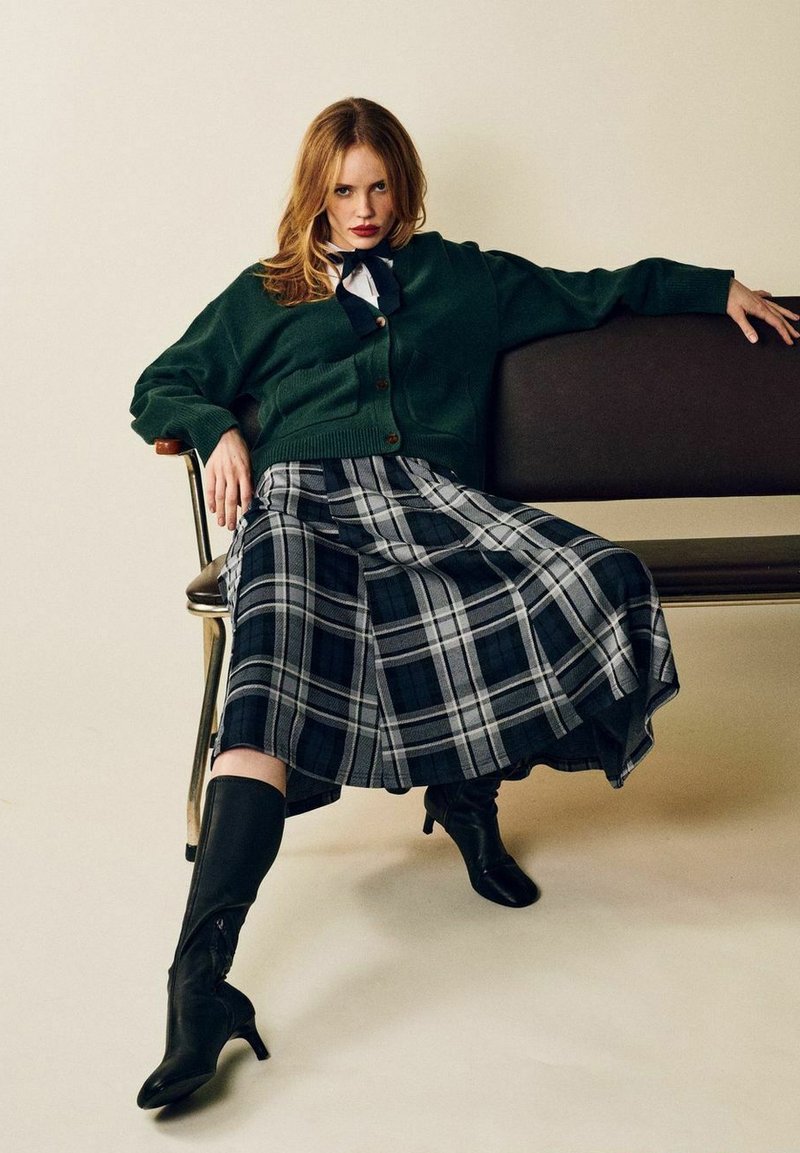 Grøn cardigan, ternet nederdel i marineblå og grå nuancer, knæhøje sorte støvler, siddende på en brun stol mod en neutral baggrund.