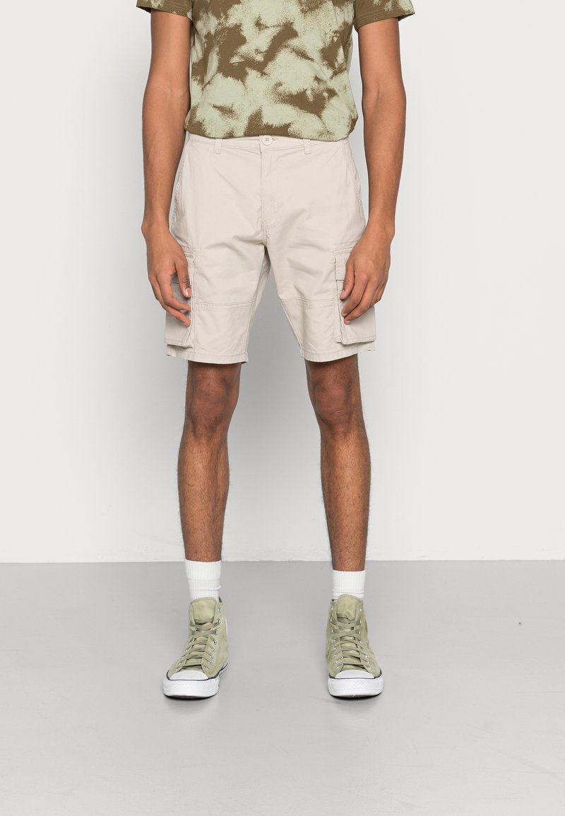 Personne portant un short cargo beige, un t-shirt tie-dye vert, des chaussettes blanches et des baskets montantes vertes, debout sur un sol gris.