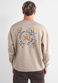 Sweat-shirt beige à manches longues, doté d'un motif floral gris et jaune au dos. Texture douce et coupe décontractée.