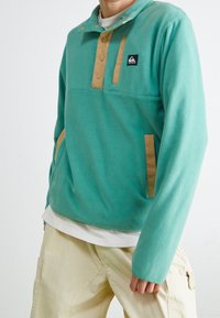 Pullover in pile color teal con dettagli in beige, dotato di un colletto alto con bottoni e due tasche frontali. Tessuto morbido, vestibilità casual.