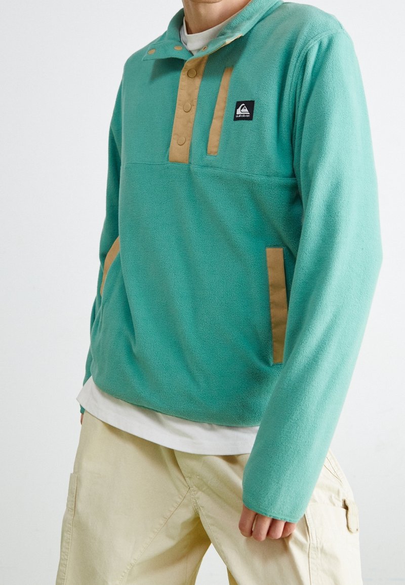 Pullover in pile color teal con dettagli in beige, dotato di un colletto alto con bottoni e due tasche frontali. Tessuto morbido, vestibilità casual.