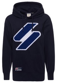 Námořnická modrá mikina s kapucí má na přední straně výrazné bílé a modré logo ve tvaru nášivky, šňůrky na kapuci a klokánkovou kapsu.