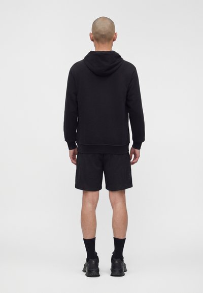 Άνδρας με ξυρισμένο κεφάλι φοράει μαύρη κουκούλα, μαύρο σορτς, μαύρες κάλτσες και μαύρα αθλητικά παπούτσια, στέκεται μπροστά σε ένα απλό ανοιχτό φόντο.