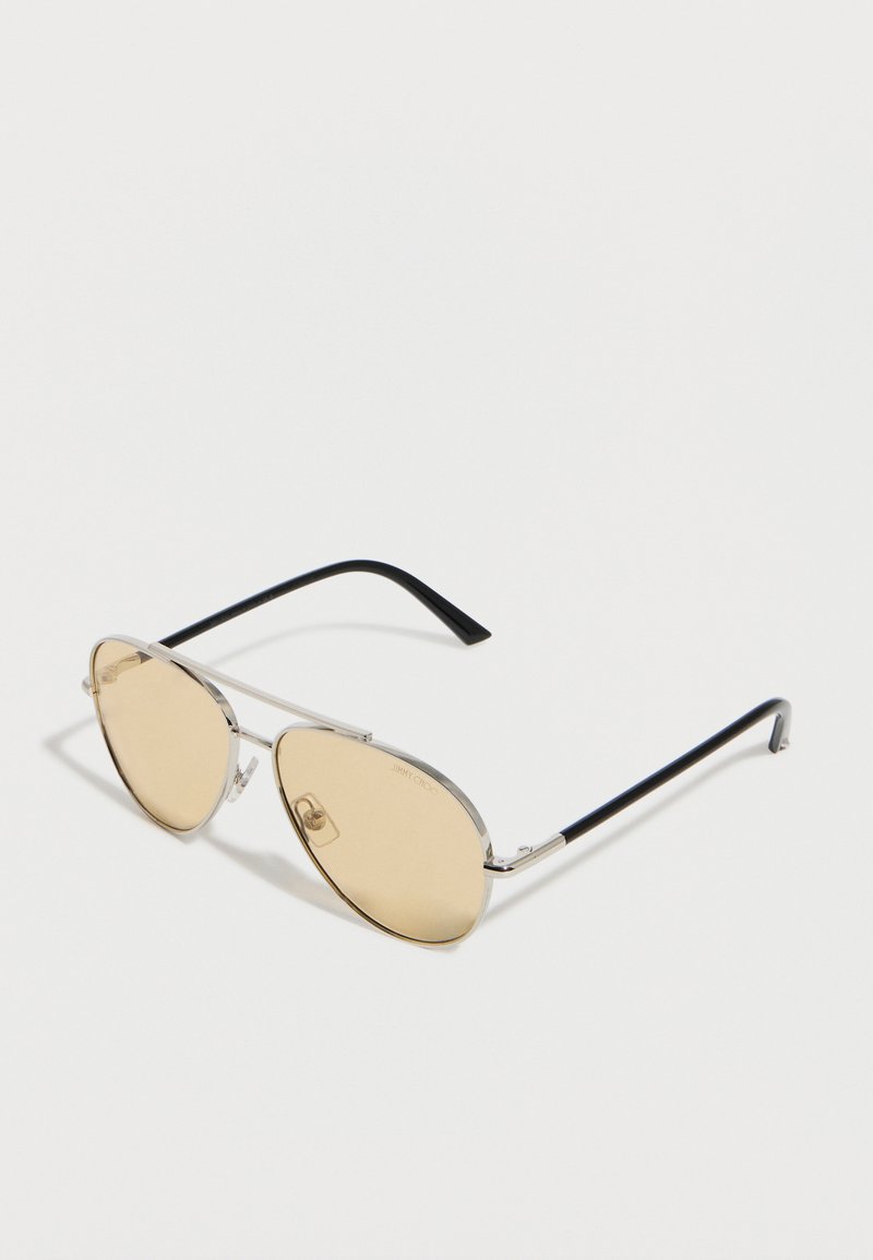 Ασημένια γυαλιά ηλίου aviator με μεταλλικό πλαίσιο, φακούς ανοιχτού καφέ χρώματος και μαύρους βραχίονες, τοποθετημένα πάνω σε λευκή επιφάνεια.