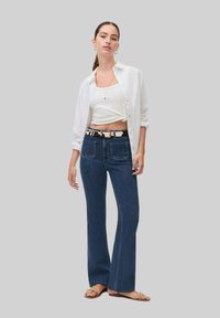 Jeans de mezclilla azul oscuro acampanados con bolsillos frontales, combinados con un top corto blanco y una camisa blanca de botonadura. Presenta un cinturón estampado.